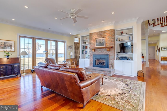 living room featuring a brick fireplace, light wood-type flooring, ornamental molding, built in features, and ceiling fan