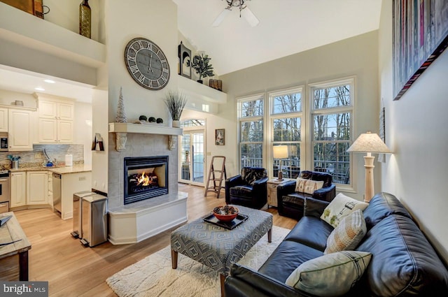 living room with a high ceiling, a tiled fireplace, ceiling fan, and light hardwood / wood-style flooring