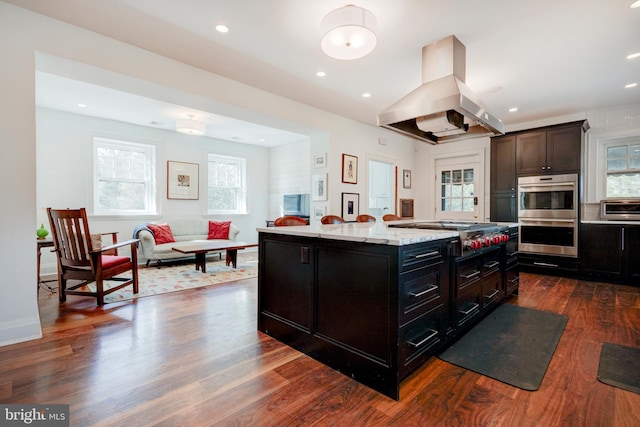 kitchen with a kitchen island, dark wood-type flooring, appliances with stainless steel finishes, island exhaust hood, and light stone counters