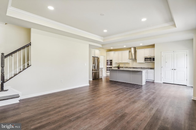 kitchen with dark wood-type flooring, a center island with sink, stainless steel appliances, wall chimney exhaust hood, and a tray ceiling