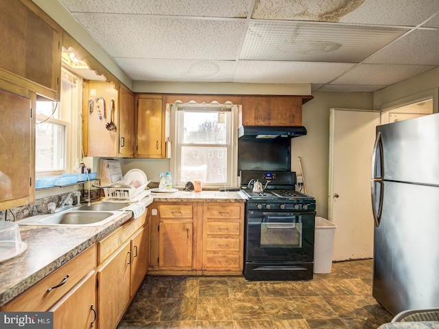 kitchen with stainless steel refrigerator, a drop ceiling, black range with gas stovetop, dark tile flooring, and sink