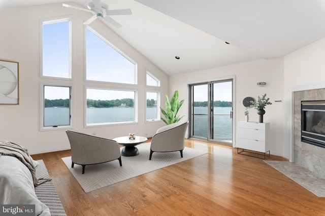 living room featuring ceiling fan, a fireplace, high vaulted ceiling, a water view, and light hardwood / wood-style flooring