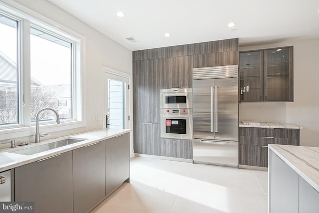 kitchen with stainless steel appliances, light tile patterned floors, sink, light stone countertops, and dark brown cabinets