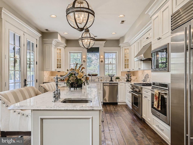 kitchen featuring light stone counters, built in appliances, pendant lighting, a notable chandelier, and wall chimney exhaust hood