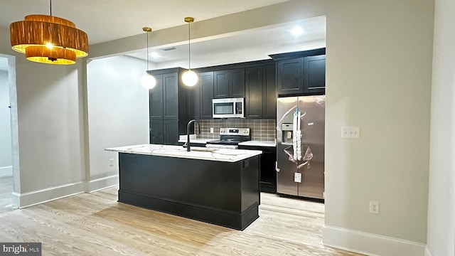 kitchen with pendant lighting, an island with sink, decorative backsplash, stainless steel appliances, and light hardwood / wood-style flooring