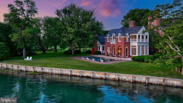 back house at dusk with a patio area, a water view, a yard, and a balcony