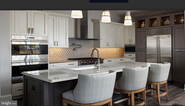 kitchen featuring an island with sink, light stone counters, hanging light fixtures, appliances with stainless steel finishes, and wall chimney range hood