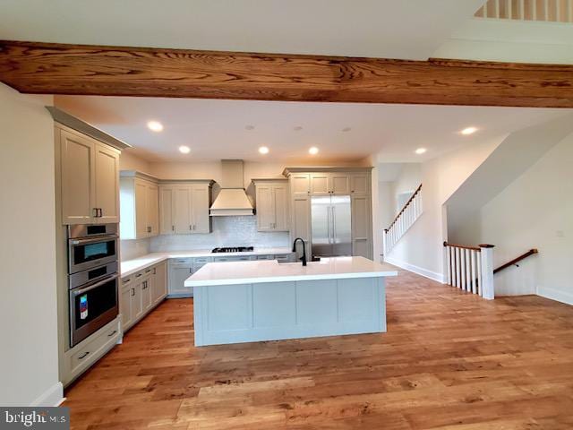 kitchen featuring light hardwood / wood-style floors, tasteful backsplash, a center island with sink, appliances with stainless steel finishes, and custom range hood