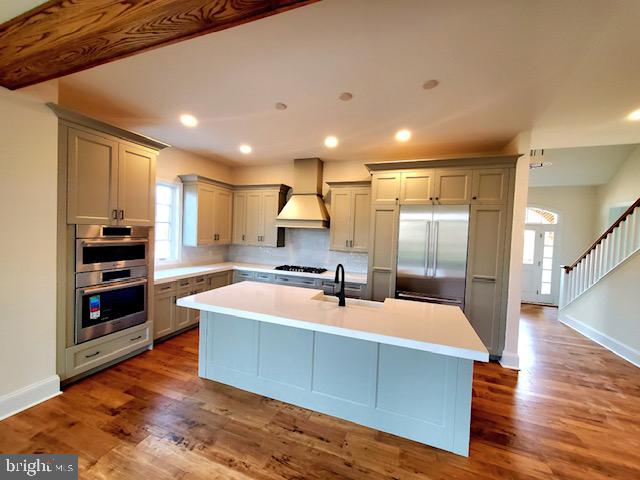 kitchen with an island with sink, custom range hood, dark wood-type flooring, and appliances with stainless steel finishes