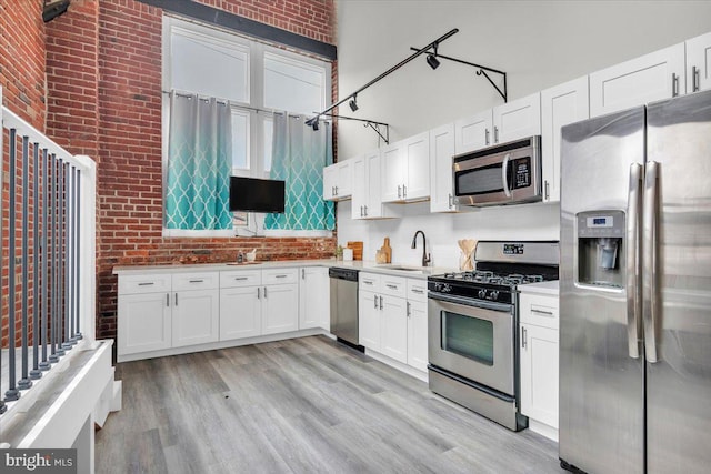 kitchen featuring brick wall, a towering ceiling, white cabinets, appliances with stainless steel finishes, and light hardwood / wood-style flooring