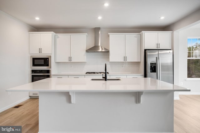 kitchen featuring appliances with stainless steel finishes, a breakfast bar, a kitchen island with sink, wall chimney range hood, and backsplash