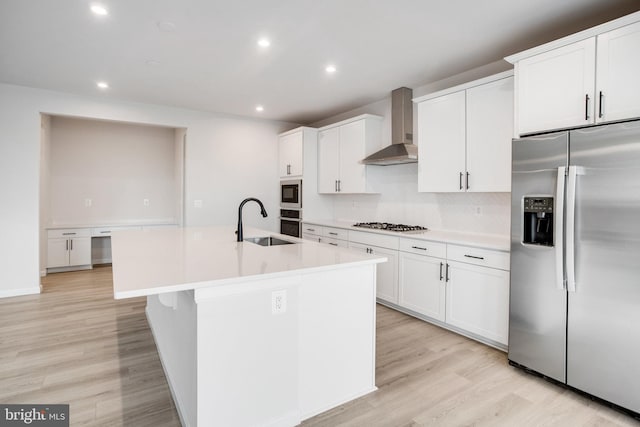 kitchen with light wood finished floors, appliances with stainless steel finishes, a kitchen island with sink, a sink, and wall chimney exhaust hood