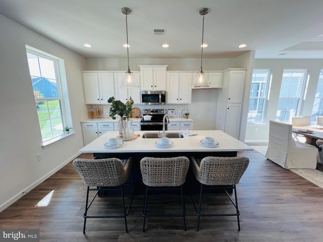 kitchen featuring dark wood-type flooring, stainless steel appliances, pendant lighting, a kitchen island with sink, and white cabinets