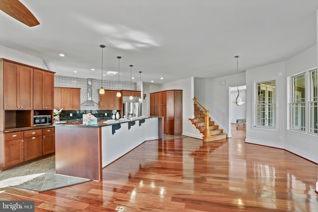 kitchen featuring appliances with stainless steel finishes, light wood-type flooring, tasteful backsplash, a kitchen island with sink, and wall chimney exhaust hood