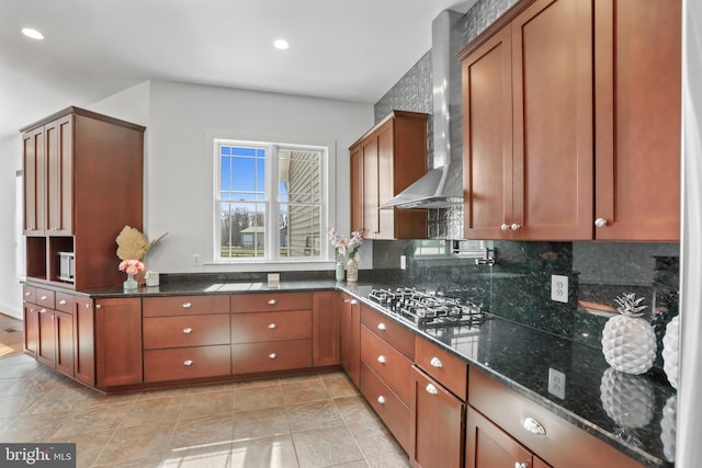 kitchen featuring appliances with stainless steel finishes, light tile floors, dark stone countertops, wall chimney range hood, and tasteful backsplash