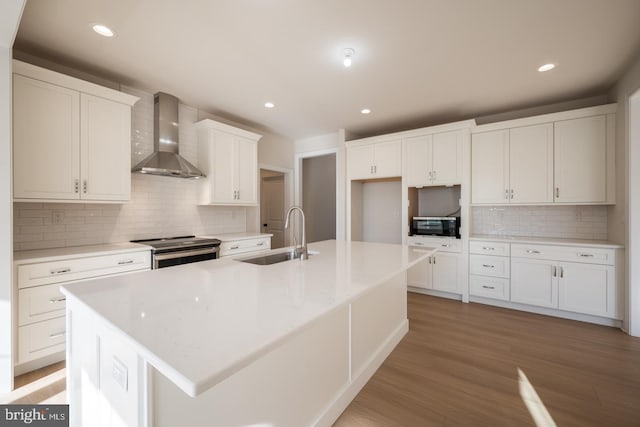 kitchen with white cabinetry, stainless steel electric range oven, a center island with sink, wall chimney exhaust hood, and sink