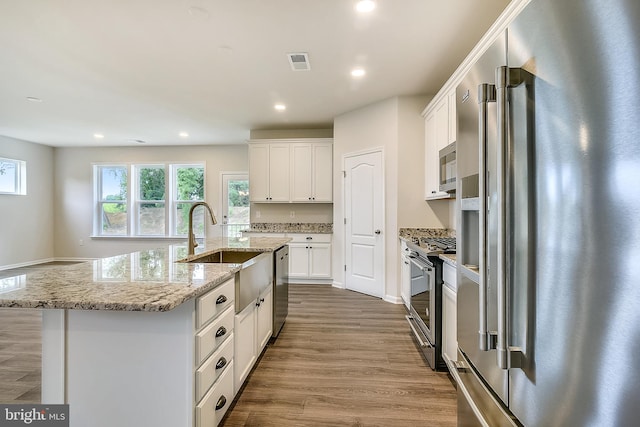 kitchen with a center island with sink, appliances with stainless steel finishes, white cabinets, light wood-type flooring, and light stone counters