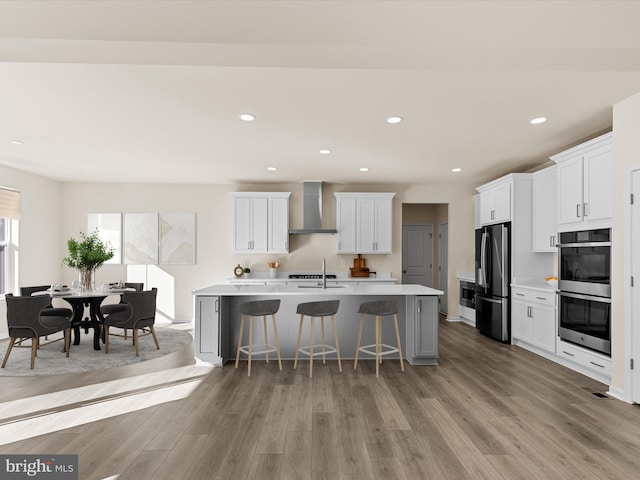 kitchen with white cabinetry, stainless steel appliances, wall chimney range hood, a center island with sink, and light wood-type flooring