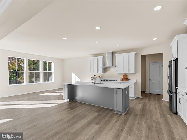 kitchen with a center island with sink, wall chimney range hood, sink, light wood-type flooring, and white cabinetry