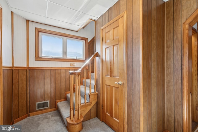 staircase featuring light colored carpet, wood walls, and a paneled ceiling