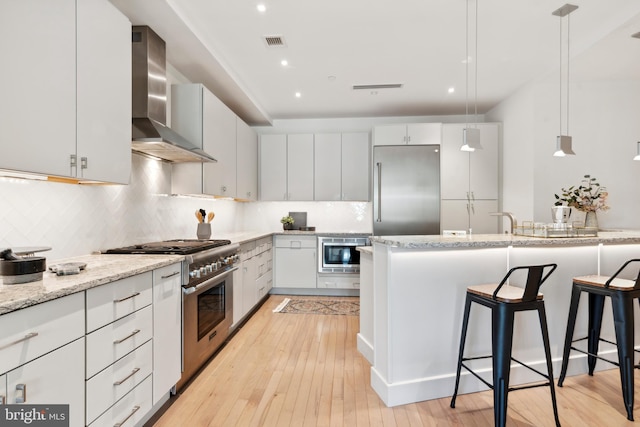 kitchen with wall chimney range hood, hanging light fixtures, white cabinetry, light hardwood / wood-style flooring, and built in appliances