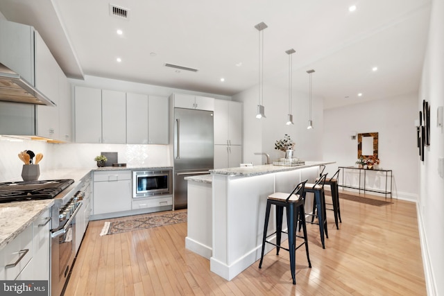 kitchen with light hardwood / wood-style floors, hanging light fixtures, built in appliances, white cabinetry, and an island with sink