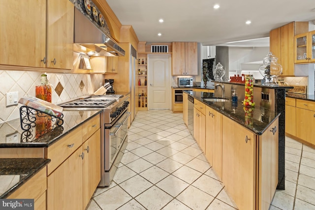 kitchen featuring sink, dark stone countertops, a center island with sink, tasteful backsplash, and high end stainless steel range