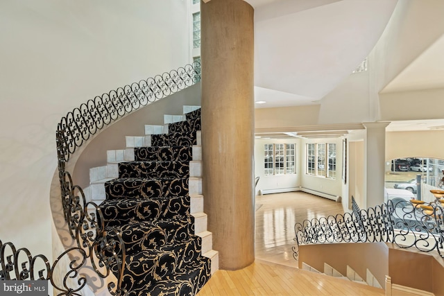 stairs featuring baseboard heating, ornate columns, light wood-type flooring, and a high ceiling