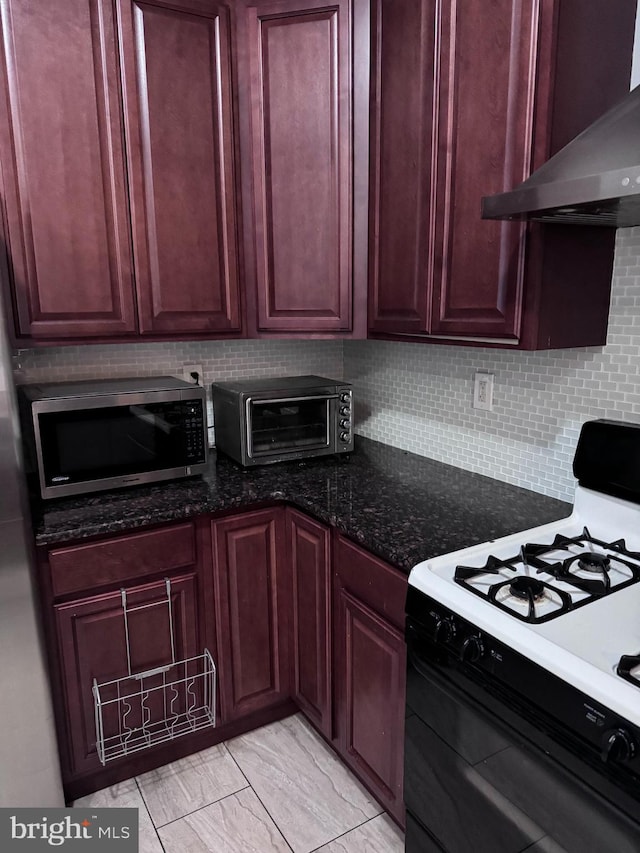 kitchen with wall chimney range hood, light tile flooring, white gas stove, tasteful backsplash, and dark stone countertops