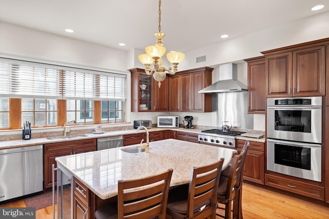 kitchen with an island with sink, stainless steel appliances, a notable chandelier, wall chimney range hood, and sink