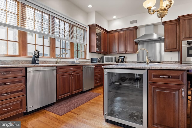 kitchen featuring a chandelier, light hardwood / wood-style floors, stainless steel appliances, wall chimney range hood, and beverage cooler