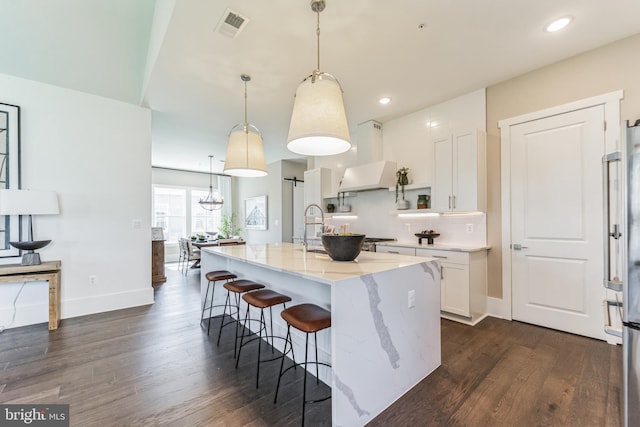 kitchen featuring a kitchen island with sink, pendant lighting, dark wood-type flooring, light stone countertops, and white cabinetry