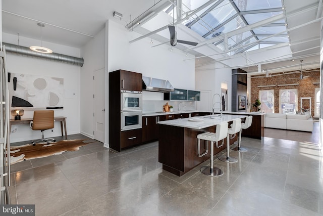 kitchen featuring dark brown cabinets, a center island with sink, high vaulted ceiling, pendant lighting, and wall chimney exhaust hood