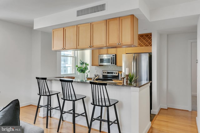 kitchen featuring dark stone counters, appliances with stainless steel finishes, light hardwood / wood-style flooring, tasteful backsplash, and a kitchen bar