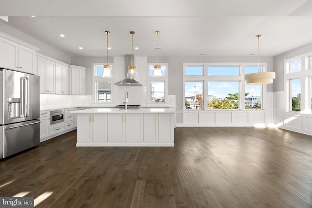 kitchen featuring high end fridge, wall chimney range hood, white cabinetry, hanging light fixtures, and an island with sink