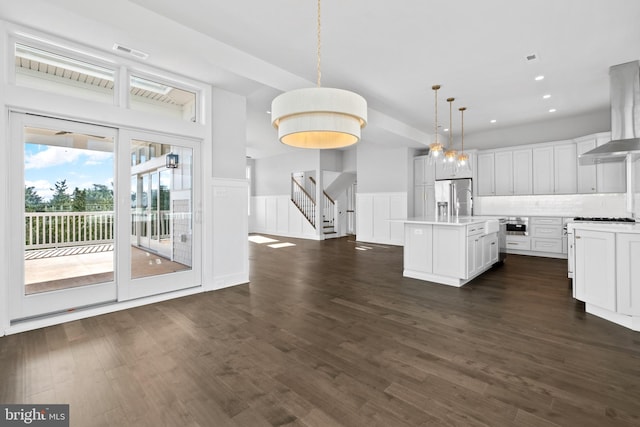 kitchen with appliances with stainless steel finishes, a kitchen island with sink, wall chimney range hood, pendant lighting, and white cabinetry