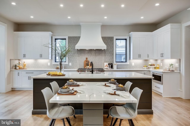 kitchen with premium range hood, stainless steel microwave, white cabinetry, a center island with sink, and light wood-type flooring