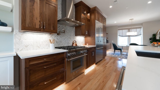 kitchen featuring wall chimney exhaust hood, backsplash, light wood-type flooring, high end appliances, and hanging light fixtures