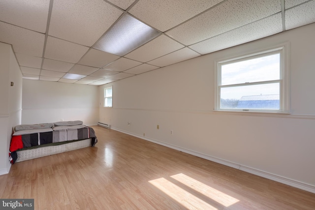 empty room with a baseboard radiator, a drop ceiling, light wood-type flooring, and a healthy amount of sunlight