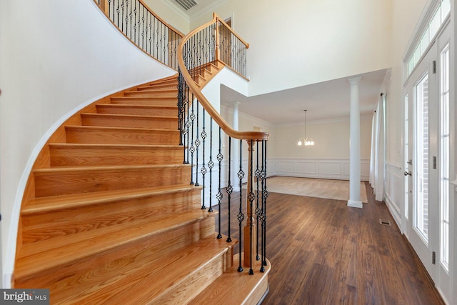 staircase with a high ceiling, decorative columns, an inviting chandelier, crown molding, and dark hardwood / wood-style flooring