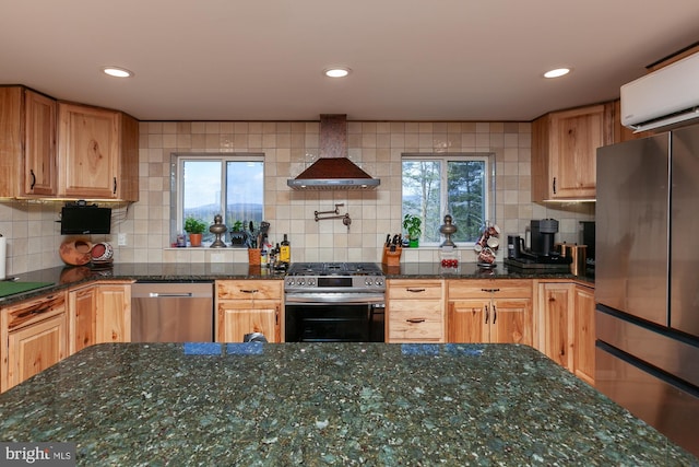 kitchen featuring dark stone countertops, a wealth of natural light, wall chimney range hood, and appliances with stainless steel finishes