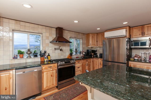 kitchen with dark stone counters, light hardwood / wood-style flooring, wall chimney exhaust hood, appliances with stainless steel finishes, and a wall unit AC