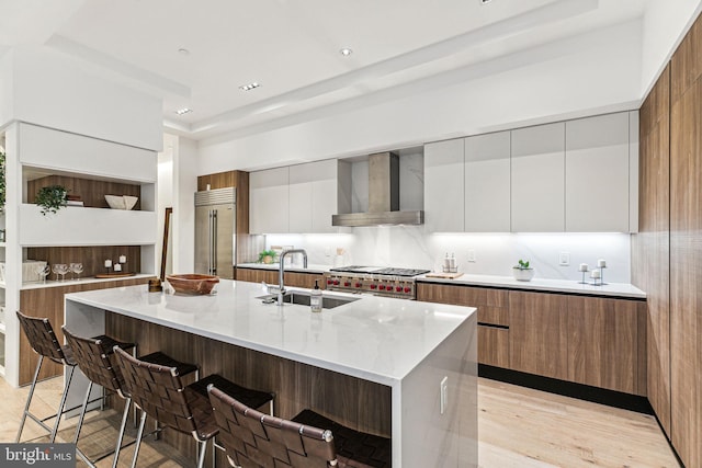 kitchen with white cabinetry, sink, wall chimney range hood, a kitchen island with sink, and stainless steel built in fridge