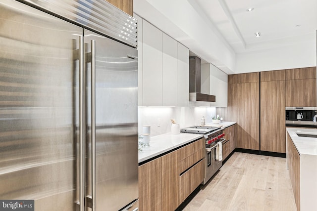 kitchen featuring wall chimney exhaust hood, premium appliances, sink, light hardwood / wood-style floors, and white cabinetry