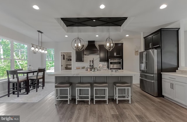 kitchen with stainless steel appliances, a tray ceiling, custom range hood, and a center island with sink