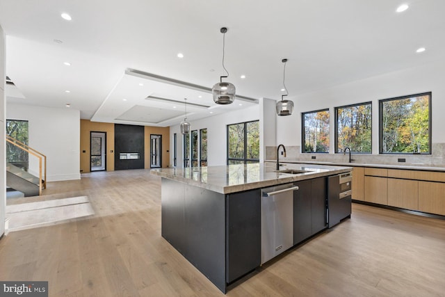 kitchen with a center island with sink, stainless steel dishwasher, light brown cabinetry, and hanging light fixtures