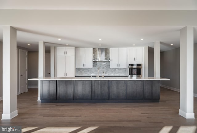 kitchen with an island with sink, stainless steel oven, dark hardwood / wood-style flooring, and wall chimney range hood