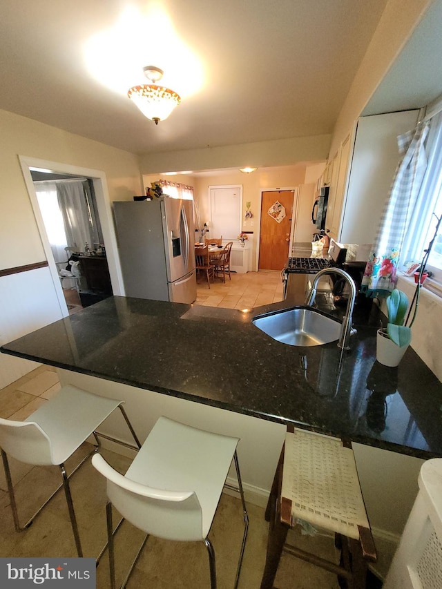 kitchen featuring light tile flooring, stove, stainless steel refrigerator with ice dispenser, dark stone countertops, and sink