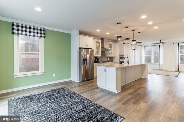 kitchen featuring decorative light fixtures, appliances with stainless steel finishes, light hardwood / wood-style floors, a kitchen island with sink, and wall chimney exhaust hood