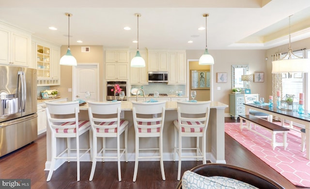 kitchen with appliances with stainless steel finishes, hanging light fixtures, light stone counters, and a kitchen island with sink